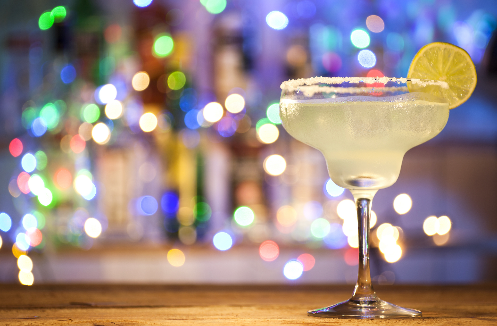 A margarita glass with a salted rim and lime slice on a bar counter, with colorful blurred lights in the background.