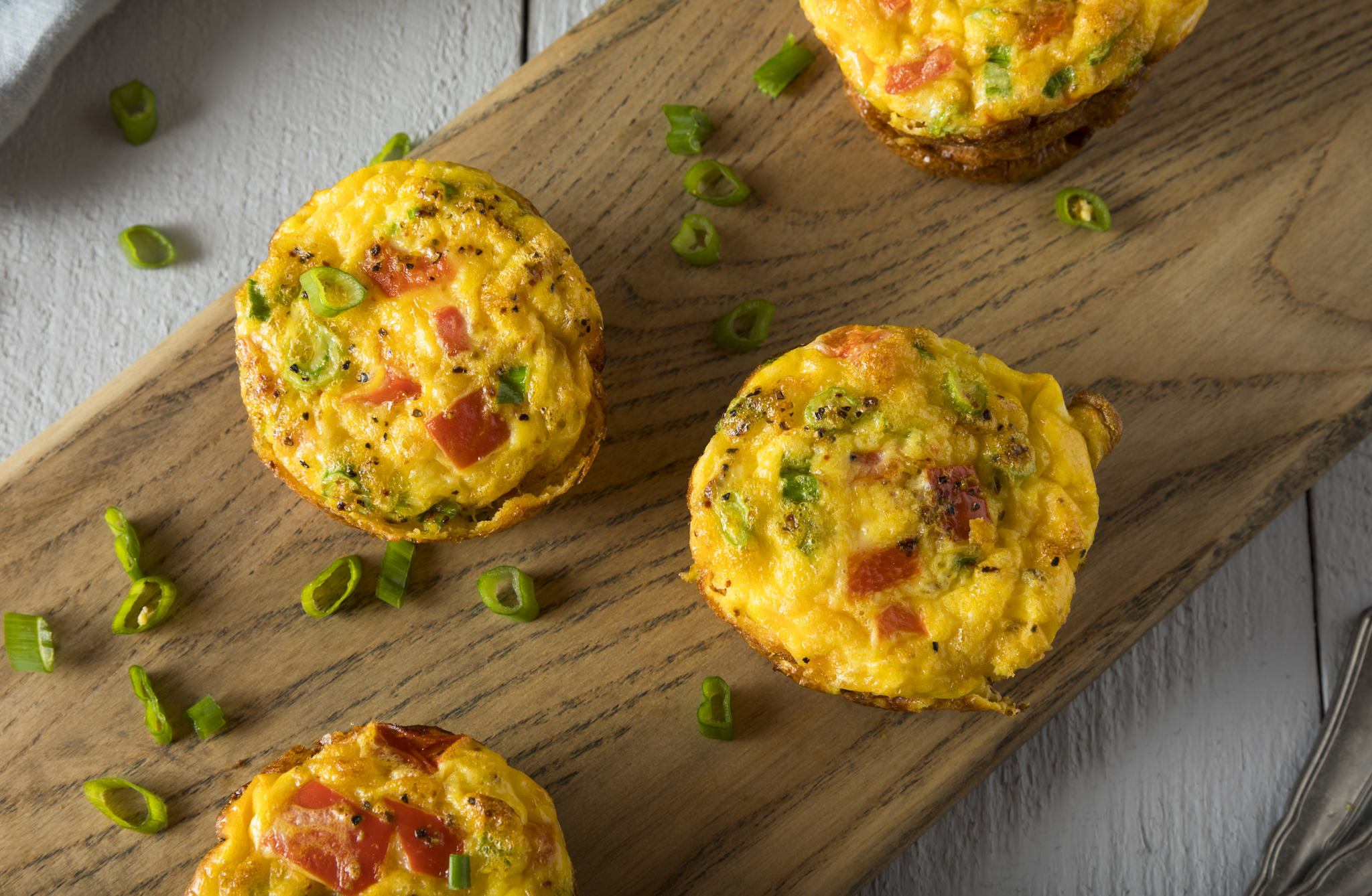 Four colorful mini frittatas with green onions and diced tomatoes are arranged on a wooden serving board, surrounded by scattered chopped green onions.