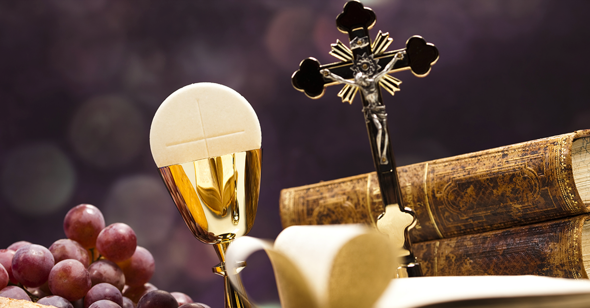 A golden chalice with a communion wafer sits beside a crucifix and old books, with clusters of grapes in the foreground, all set against a softly blurred background.