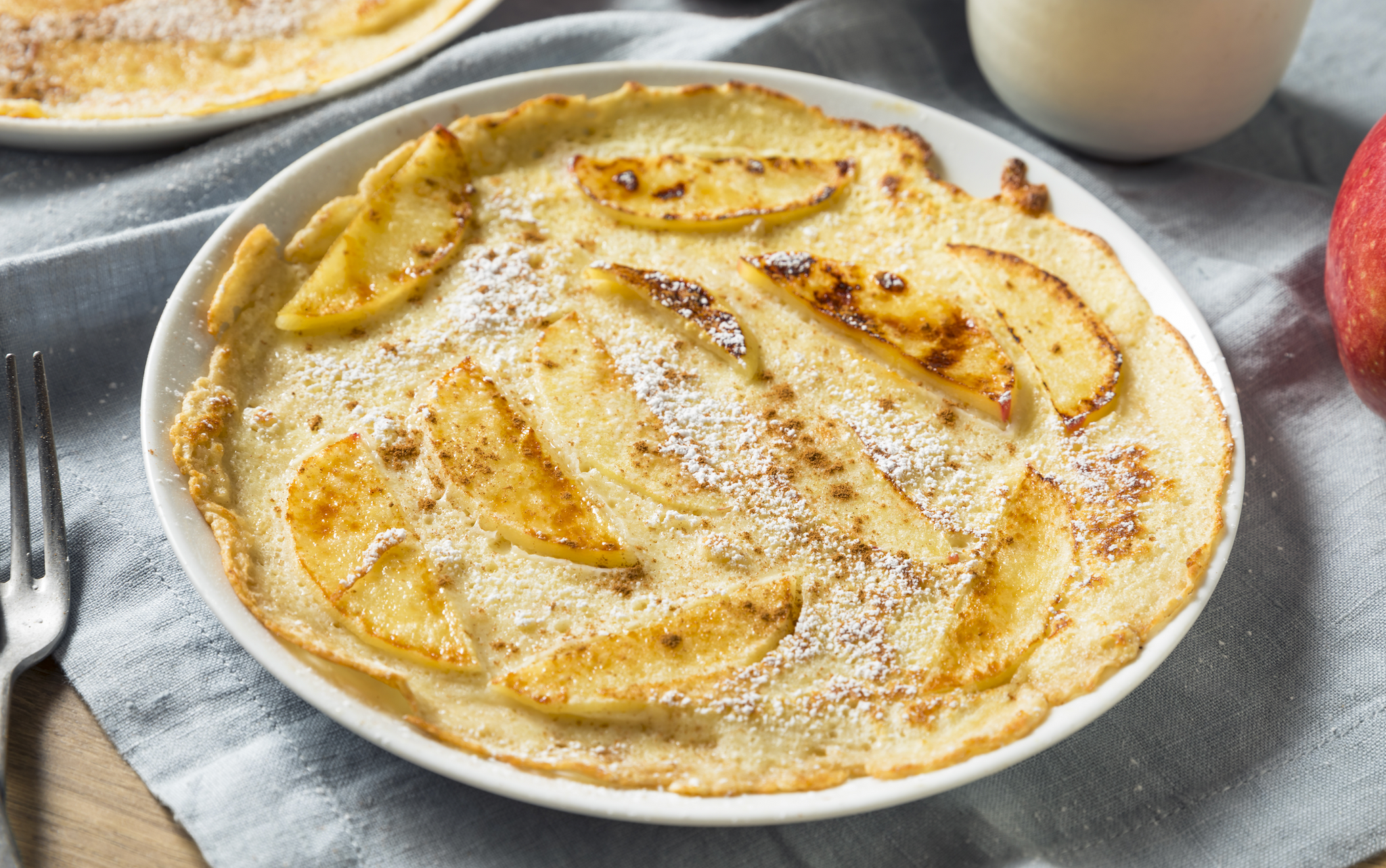 A round plate holds a golden-brown apple pancake topped with powdered sugar, surrounded by a fork, a red apple, and a glass of milk on a textured gray cloth.