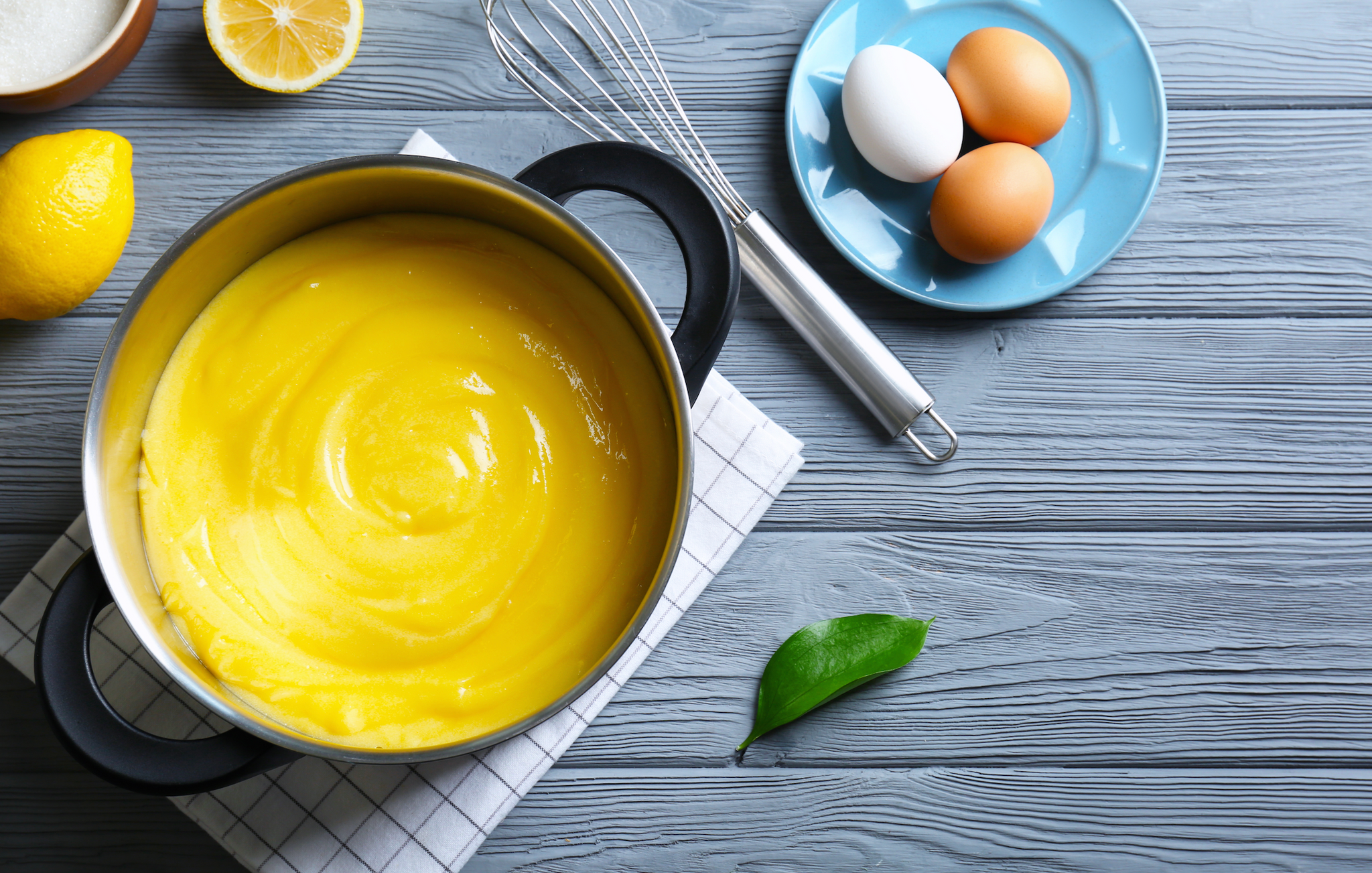 Pot of bright yellow lemon curd with lemons, eggs, a whisk, and a napkin on a gray wooden surface.