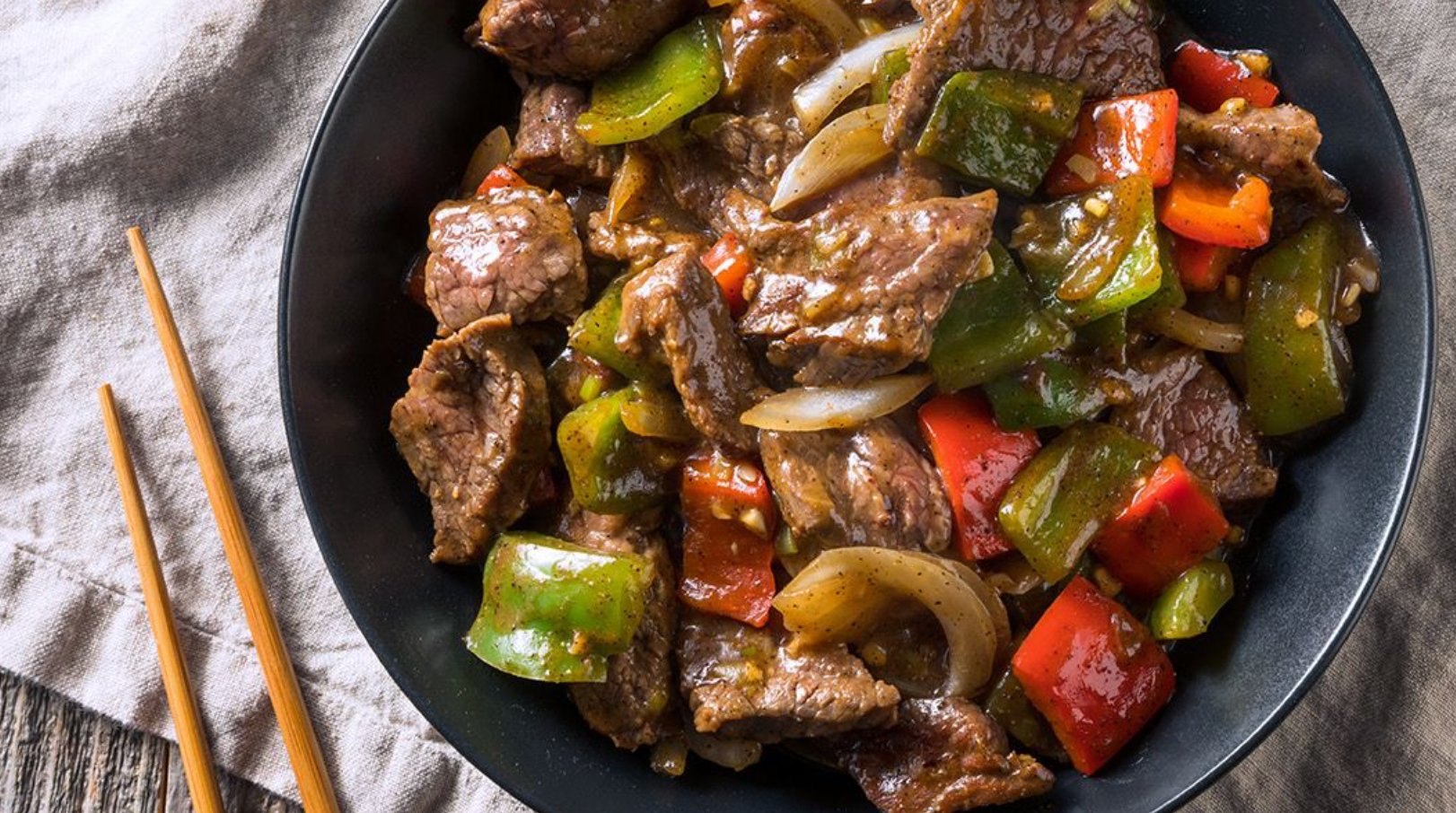 Bowl of beef stir-fry with bell peppers and onions in a glossy sauce, served beside a pair of chopsticks.