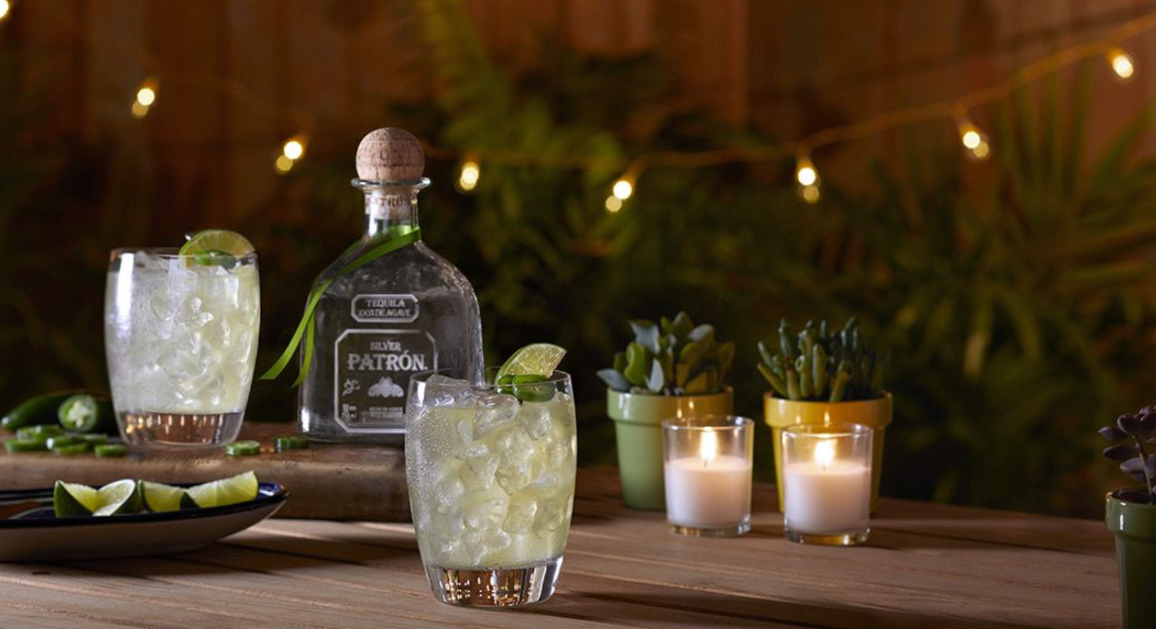 Tequila bottle with two iced lime cocktails on a wooden table, surrounded by candles, plants, and string lights.