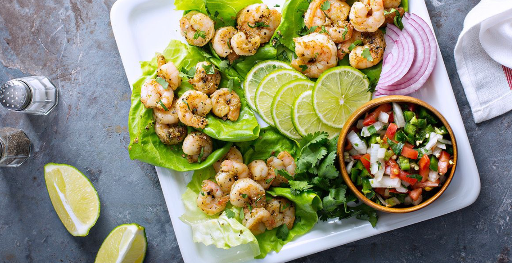 A platter of seasoned shrimp on lettuce leaves is arranged with lime slices, red onion, and a bowl of fresh salsa, set against a textured gray background.