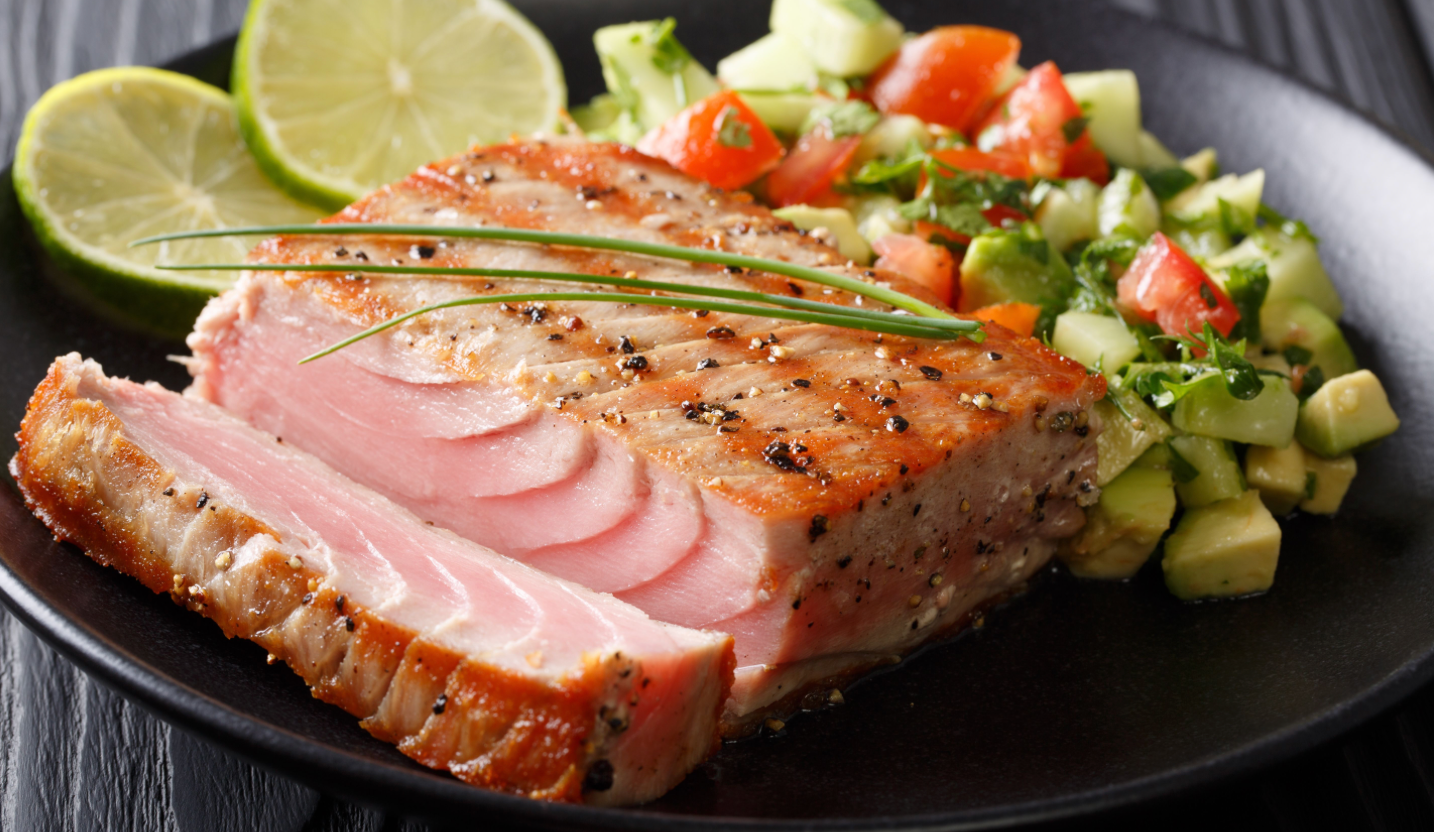 Seared tuna steak served with lime slices and a fresh avocado and tomato salad on a black plate.