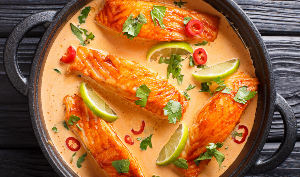 Three pieces of cooked salmon are arranged in a creamy sauce, garnished with lime wedges, cilantro, and sliced red chilies, in a black pot on a textured wooden surface.