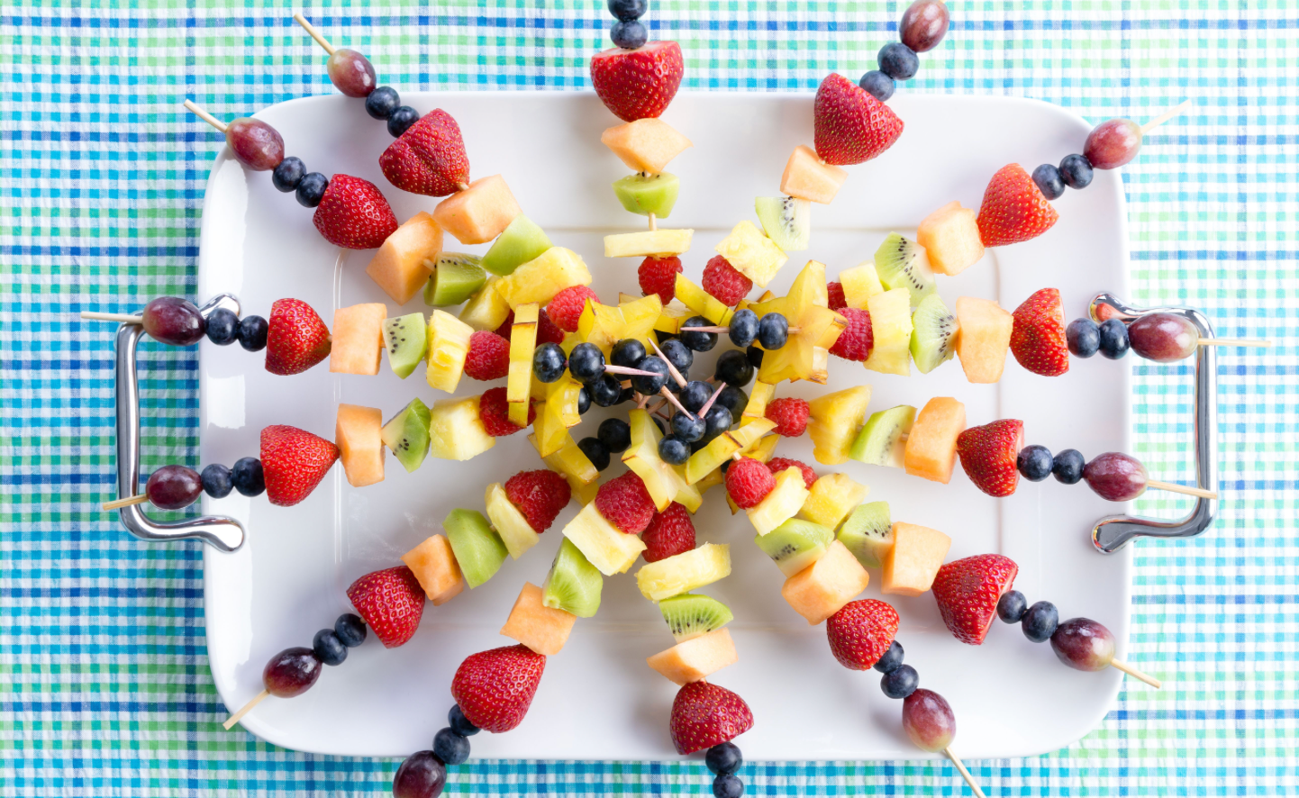A platter of colorful fruit skewers, featuring strawberries, blueberries, kiwi, cantaloupe, and pineapple, arranged in a starburst pattern on a checkered blue and green tablecloth.