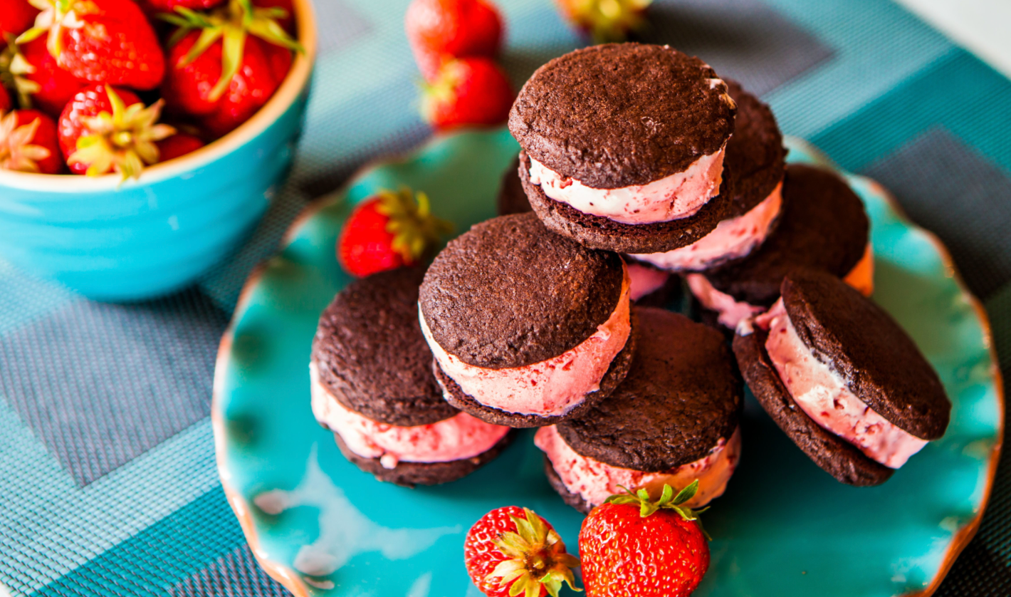 Chocolate cookie sandwiches filled with strawberry ice cream on a turquoise plate with fresh strawberries.