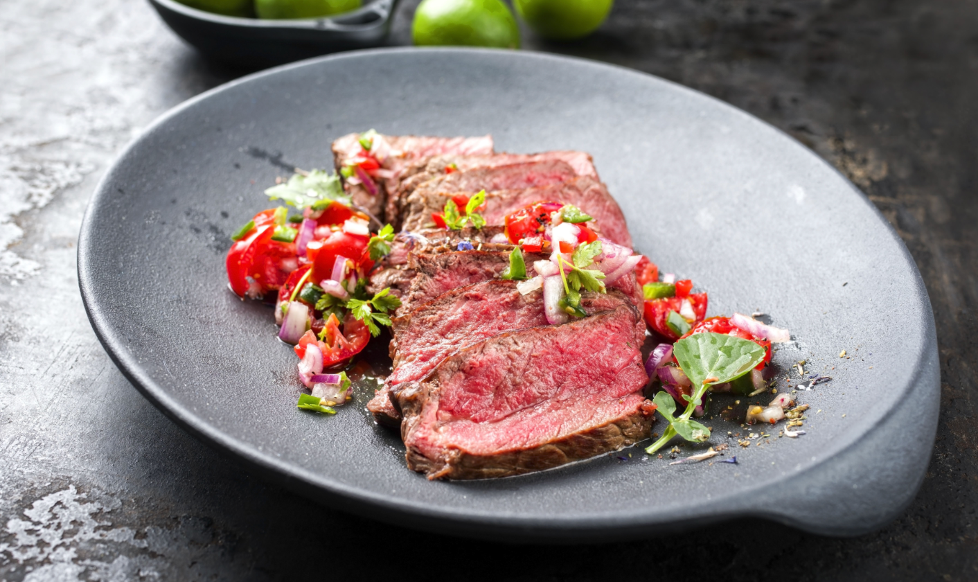 Sliced beef rests on a dark plate, topped with a vibrant mix of diced tomatoes, onions, and cilantro, with fresh limes in the background, suggesting a flavorful, fresh meal.
