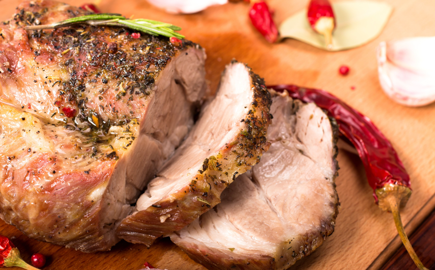 Sliced roasted meat seasoned with herbs and spices, shown juicy and tender on a wooden board with dried chilies and garlic nearby.