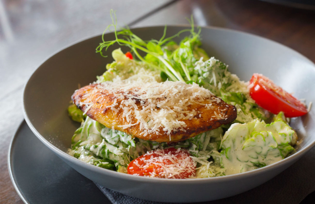 Caesar style salad with grilled chicken, cherry tomatoes, fresh greens, and grated cheese in a grey bowl