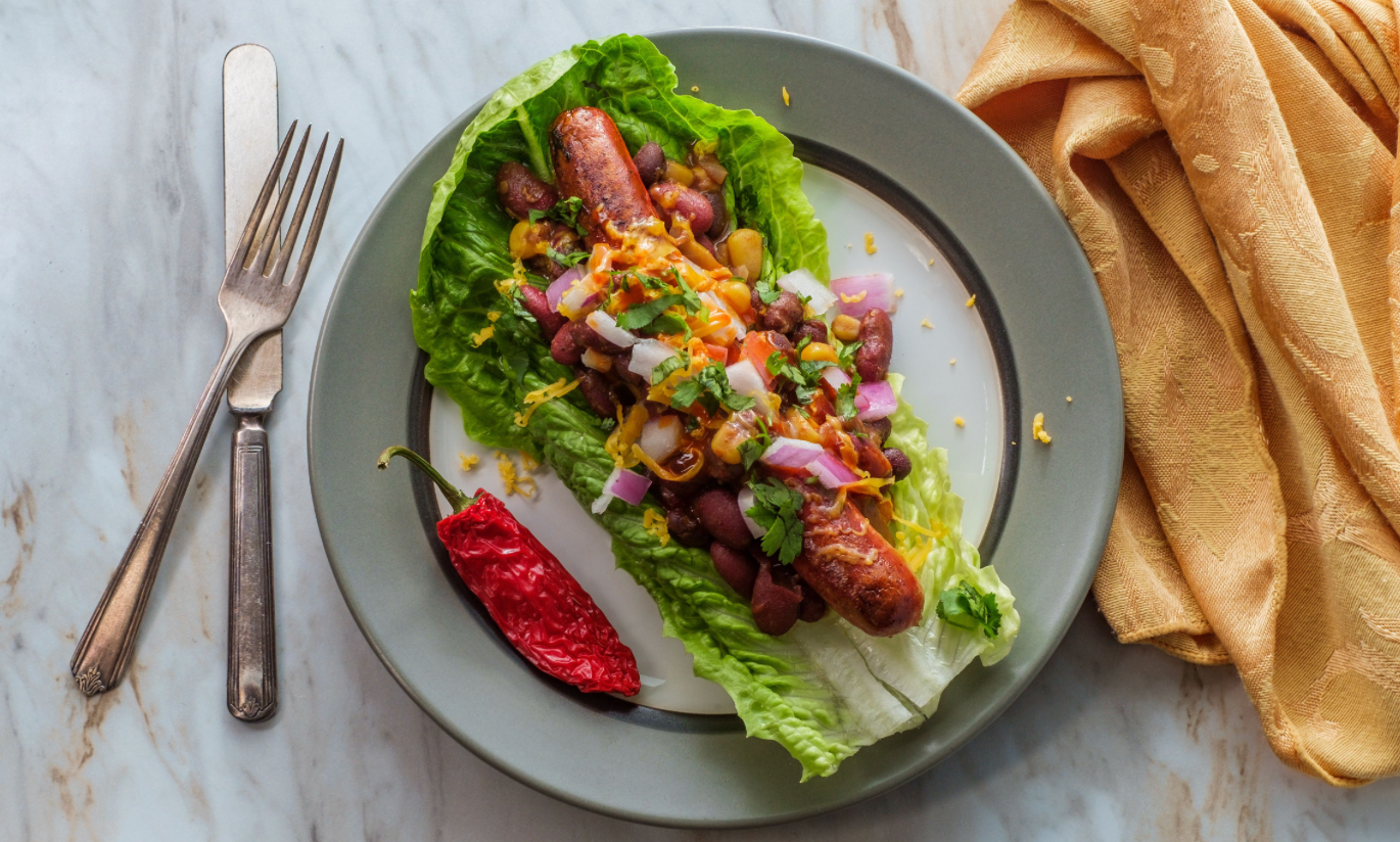 Lettuce-wrapped sausage topped with beans, onions, cheese, and herbs, served with a red pepper on a plate.