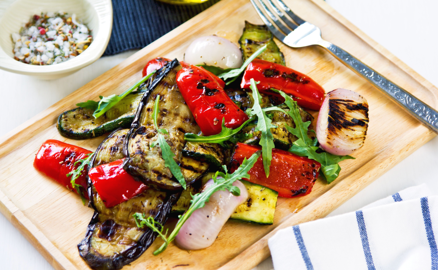 Grilled vegetables including peppers, eggplant, zucchini and onions served with arugula on a wooden board.
