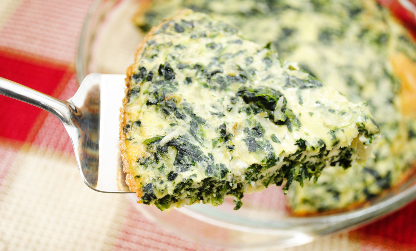 A slice of creamy spinach quiche being lifted with a metal spatula from a glass pie dish.