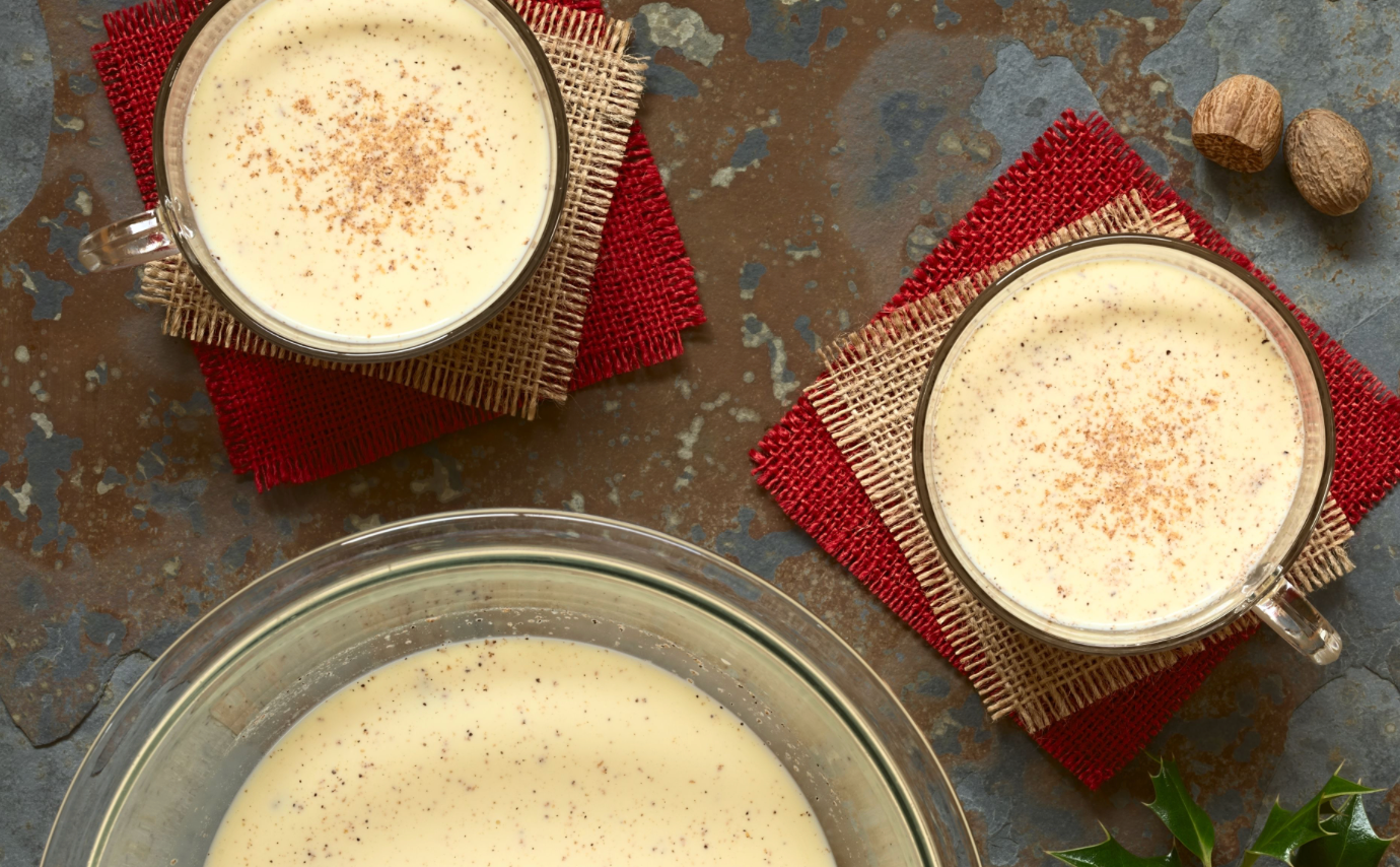 Glass mugs of creamy eggnog topped with nutmeg on red coasters beside a large bowl and whole nutmeg.