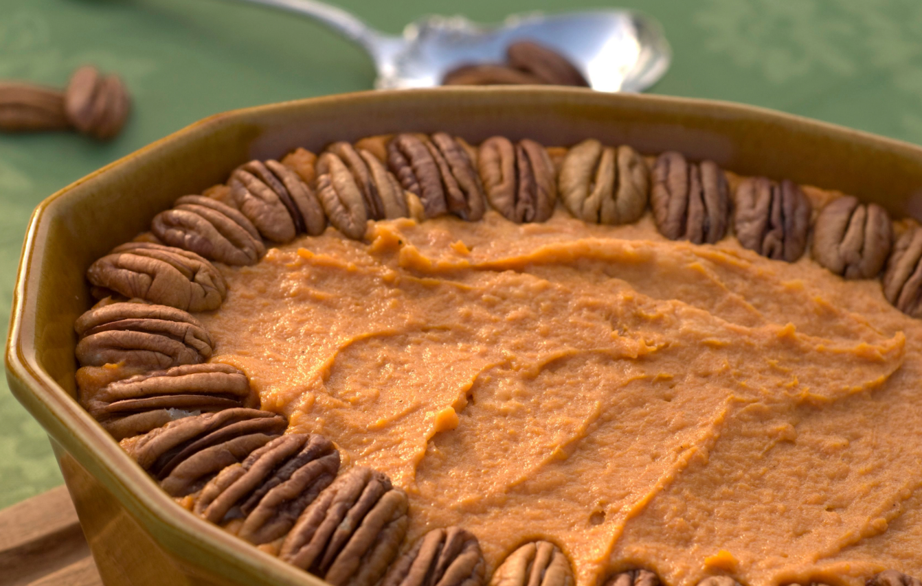 Sweet potato casserole in a brown dish topped with a ring of pecans, with a spoon and extra pecans in the background.