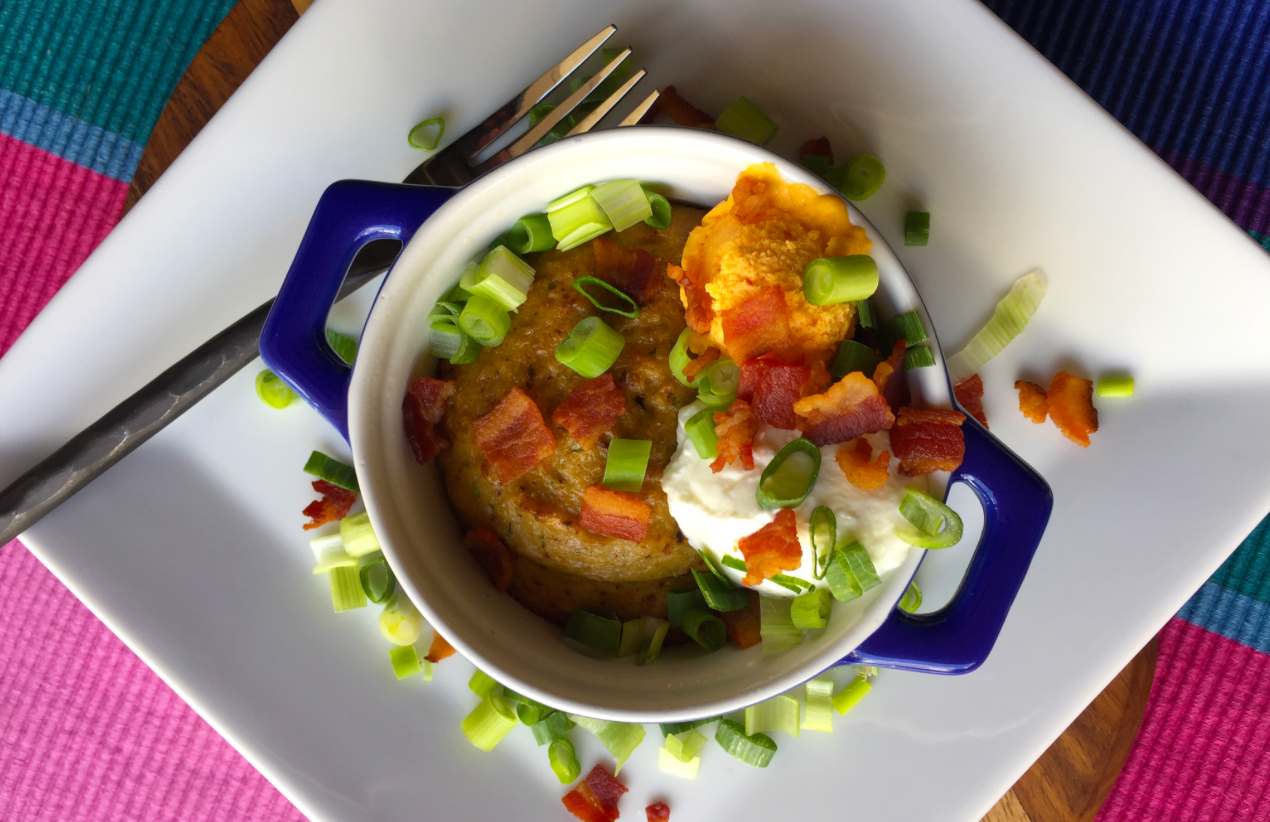 Small dish of savory baked batter topped with sour cream, bacon bits, and chopped green onions on a white plate.