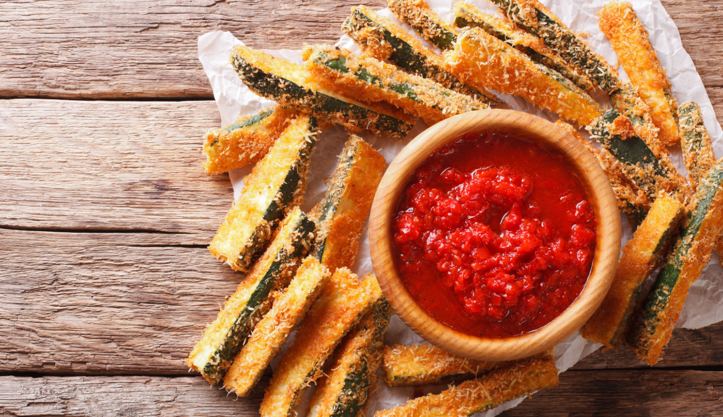 Crispy, golden zucchini sticks are arranged around a wooden bowl filled with rich marinara sauce, set on a rustic wooden surface.