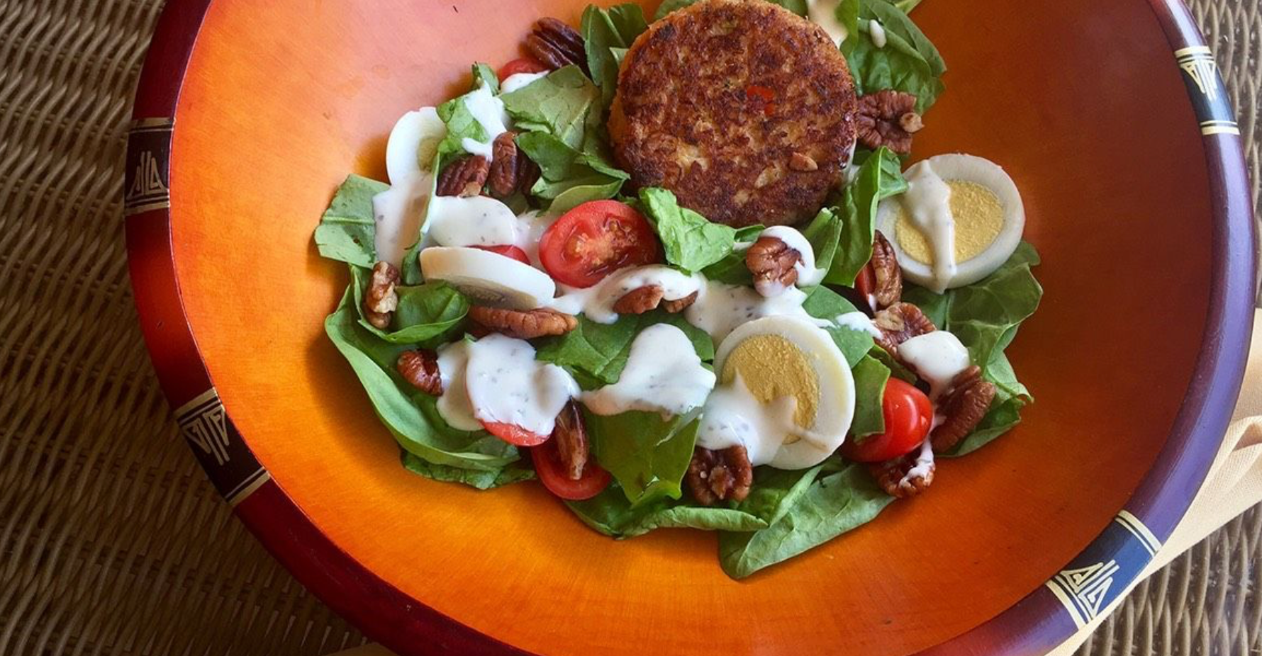 Spinach salad with tomatoes, pecans, sliced eggs, creamy dressing, and a crispy patty served in a wooden bowl.