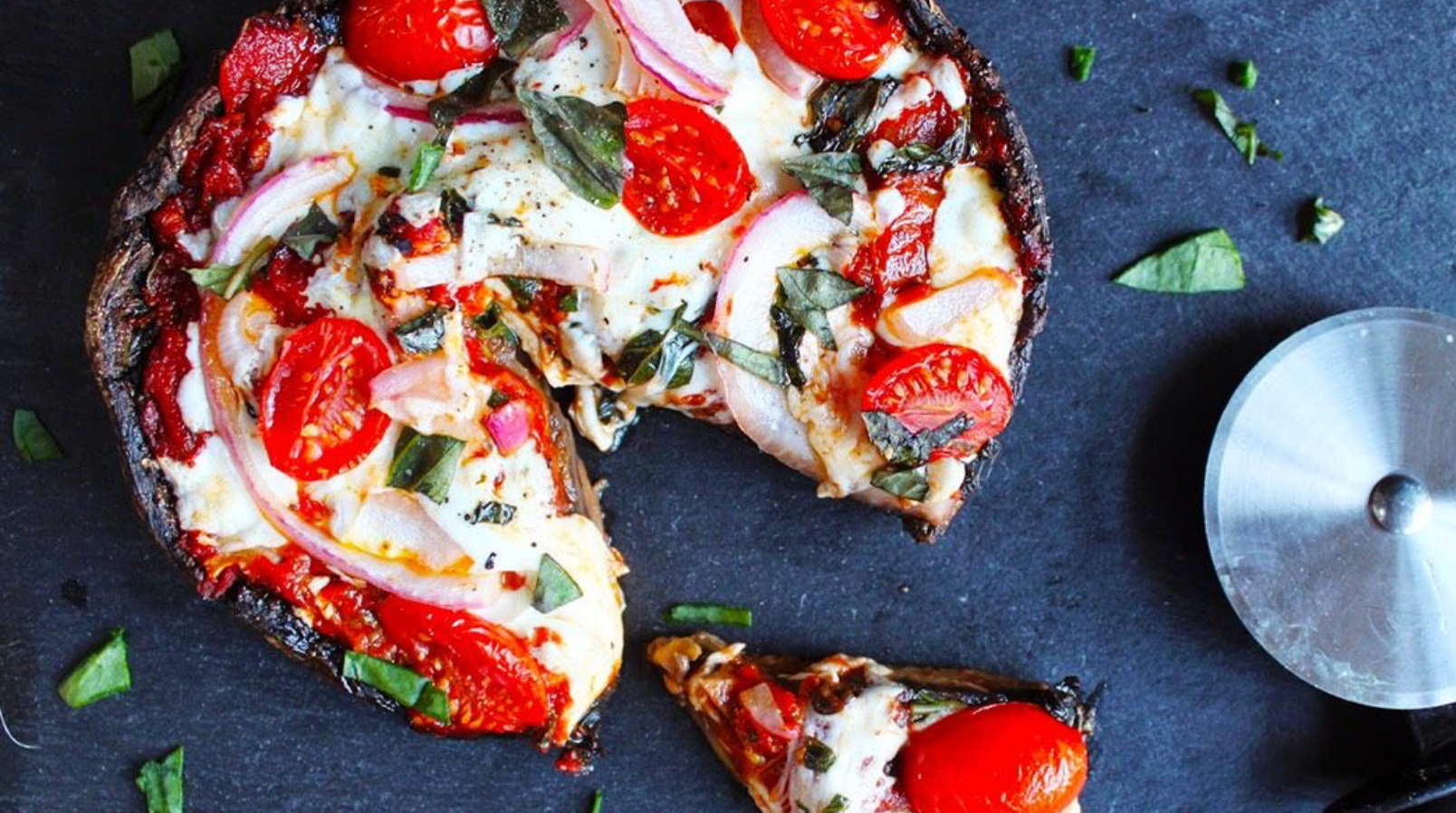 Personal sized pizza topped with tomatoes, onions, basil, and melted cheese, with a slice cut and a pizza cutter nearby.