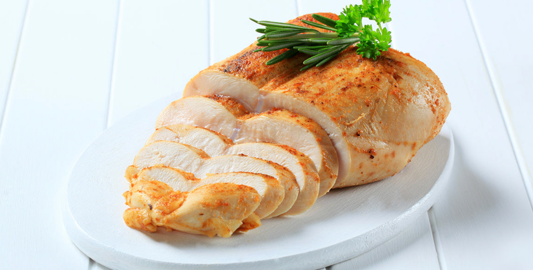 Sliced cooked chicken breast is arranged on a white plate, garnished with fresh herbs, showcasing a golden-brown crust and tender, juicy meat on a light wooden surface.