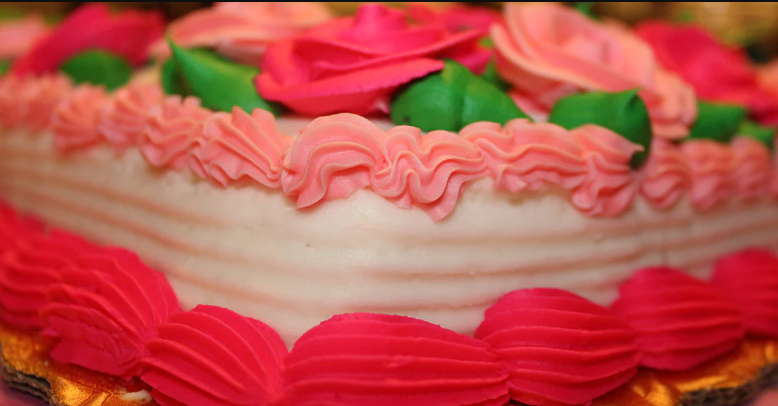 A beautifully decorated cake features pink and green frosting, with intricate ruffles and rose-shaped designs, set against a blurred background.