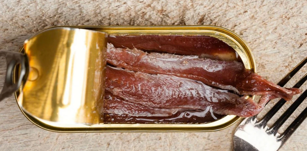 A partially opened gold tin reveals several fillets of anchovies in oil, resting on a wooden surface next to a fork.