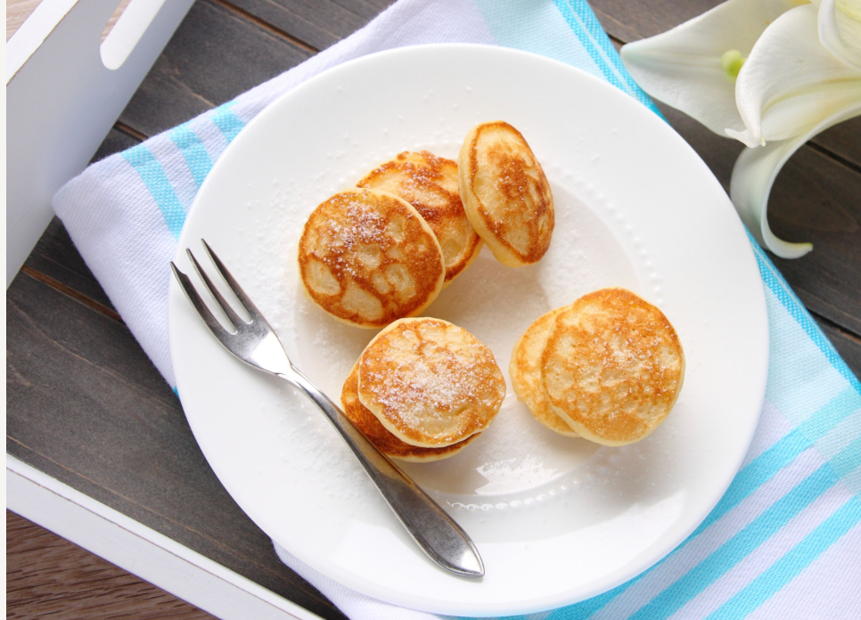 A white plate holds five golden-brown, fluffy pancakes dusted with powdered sugar, accompanied by a fork, set on a blue-striped towel next to a wooden surface and a white flower.