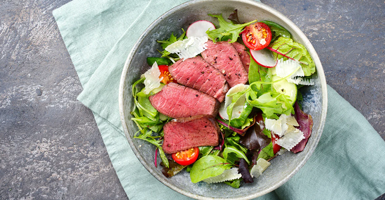 A bowl of mixed greens topped with slices of pink roast beef, cherry tomatoes, radishes, and shaved cheese, set on a light green cloth against a textured gray background.