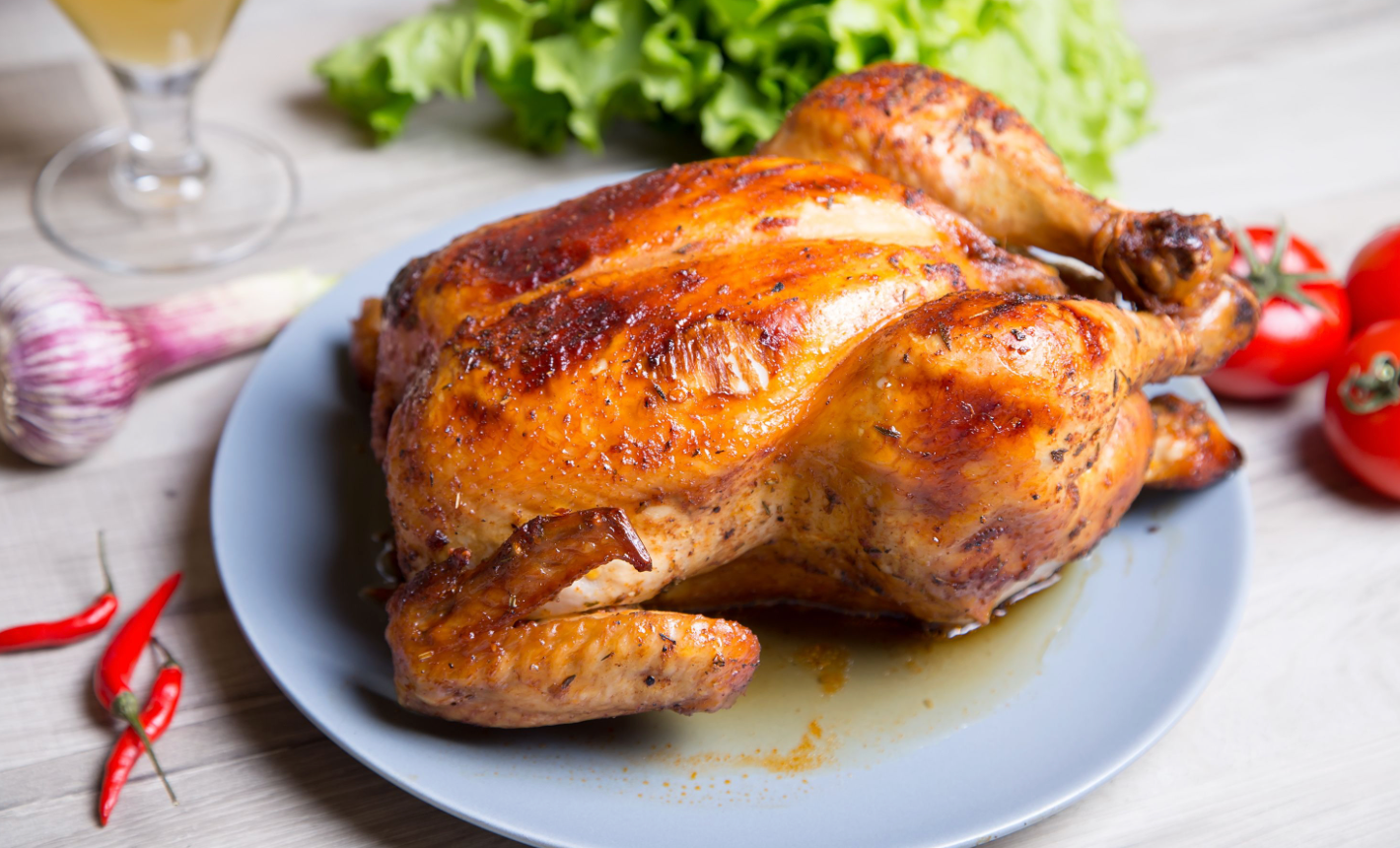 Golden roasted whole chicken on a plate with tomatoes, garlic, chili peppers, and lettuce in the background.