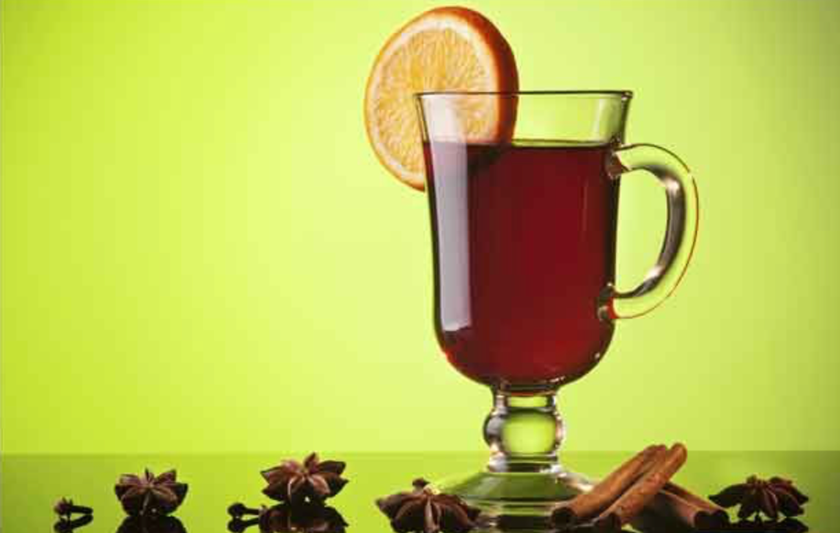 Hot spiced drink in a glass mug with an orange slice, surrounded by star anise and cinnamon sticks.