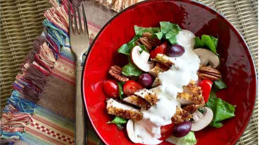 Crispy chicken salad with lettuce, tomatoes, mushrooms, pecans, grapes, and creamy dressing in a red bowl.