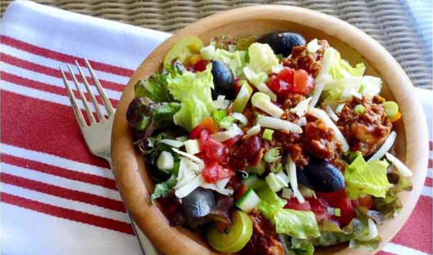 A wooden bowl filled with a colorful salad featuring lettuce, tomatoes, olives, cheese, and green onions sits on a striped red and white napkin next to a silver fork.