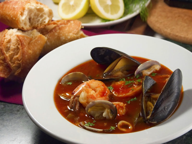 A bowl of seafood stew with shrimp, clams, and mussels sits on a table, garnished with parsley, alongside crusty bread and slices of lemon in the background.