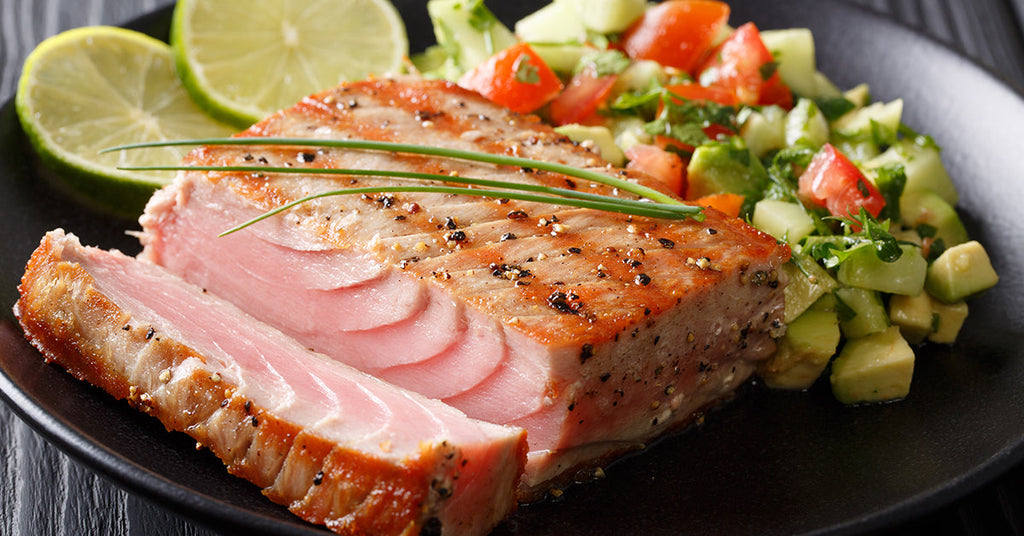 Seared tuna steak sliced to show its pink center, served with avocado-tomato salad and lime slices on a black plate.