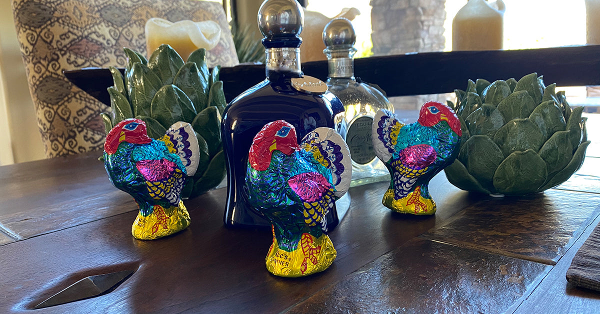 Three colorful, foil-wrapped turkey decorations are placed on a wooden table, accompanied by decorative bottles and a green artichoke centerpiece, creating a festive Thanksgiving atmosphere.