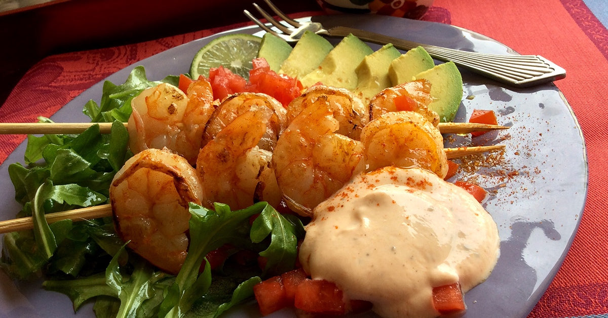 Grilled shrimp skewers with lettuce, diced tomatoes, avocado slices, lime, and a creamy dipping sauce on a plate.