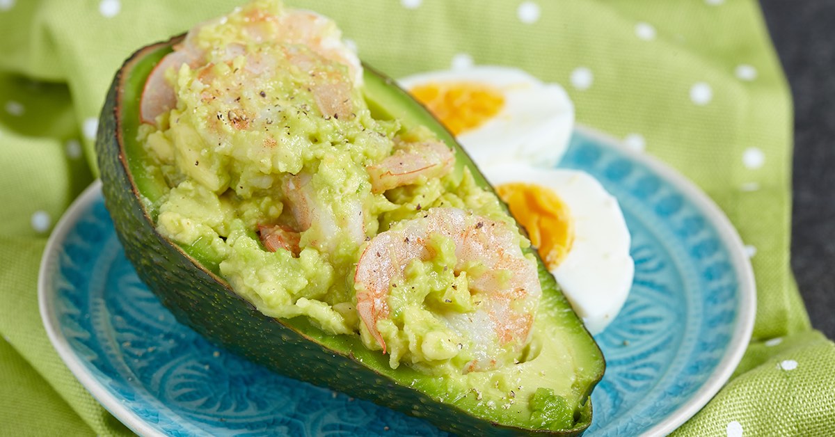A halved avocado filled with mashed avocado and shrimp sits on a blue plate next to two halved boiled eggs, with a green polka-dotted cloth in the background.