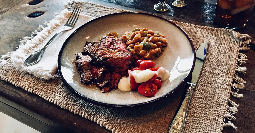 Plate of sliced steak with beans and cherry tomatoes topped with cream, set on a rustic table with cutlery.