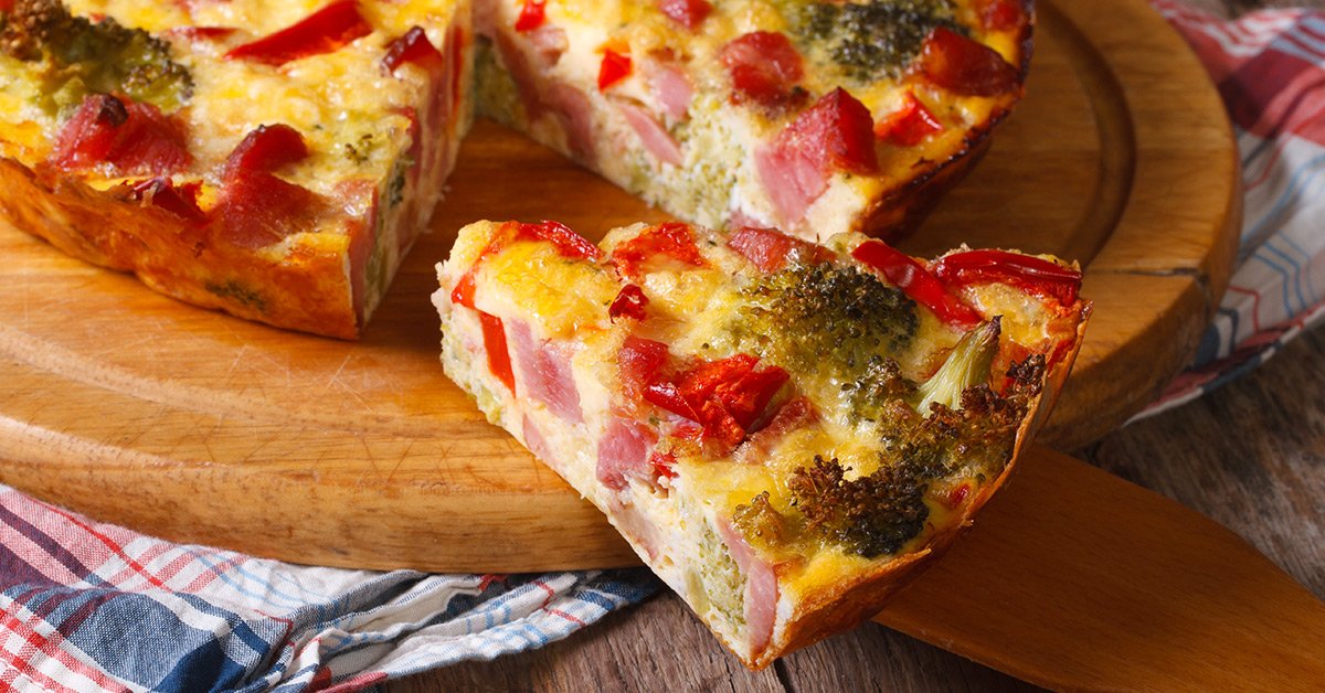 Slice of baked vegetable and ham quiche on a wooden board.