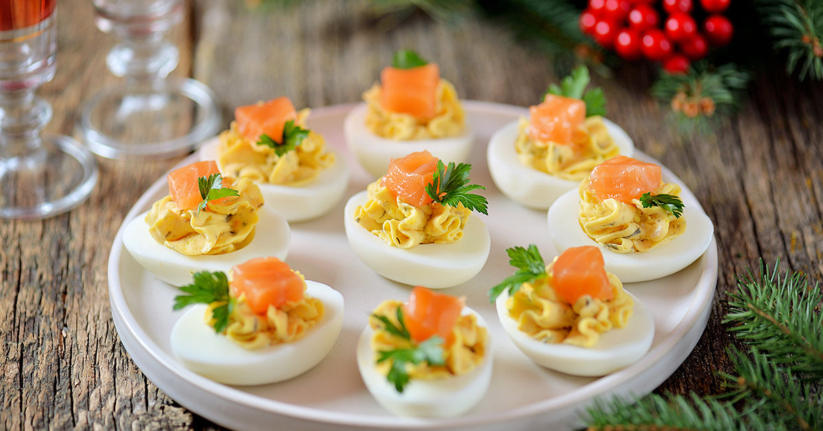 A white plate holds several deviled eggs topped with yellow filling, smoked salmon, and parsley, set on a rustic wooden table with festive greenery and red berries in the background.