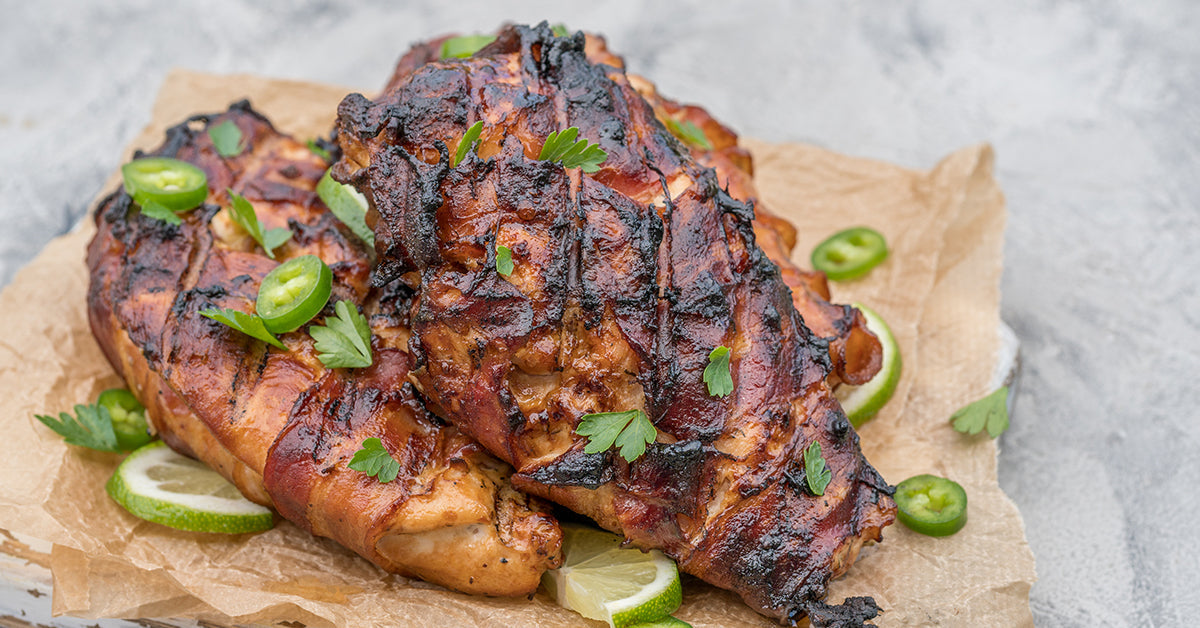 Two grilled chicken breasts wrapped in bacon, garnished with fresh parsley and sliced jalapeños, are resting on a piece of parchment paper with lime wedges nearby.