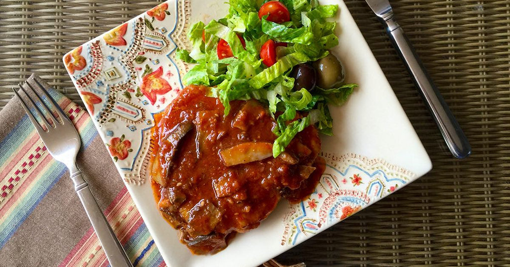 Plate with tomato-sauced meat and veggies, served with a fresh green salad and olives on a patterned dish.