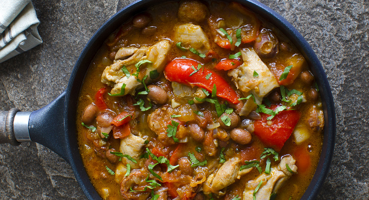 Pan of hearty stew with chicken, beans, red peppers, and fresh herbs in a rich broth.