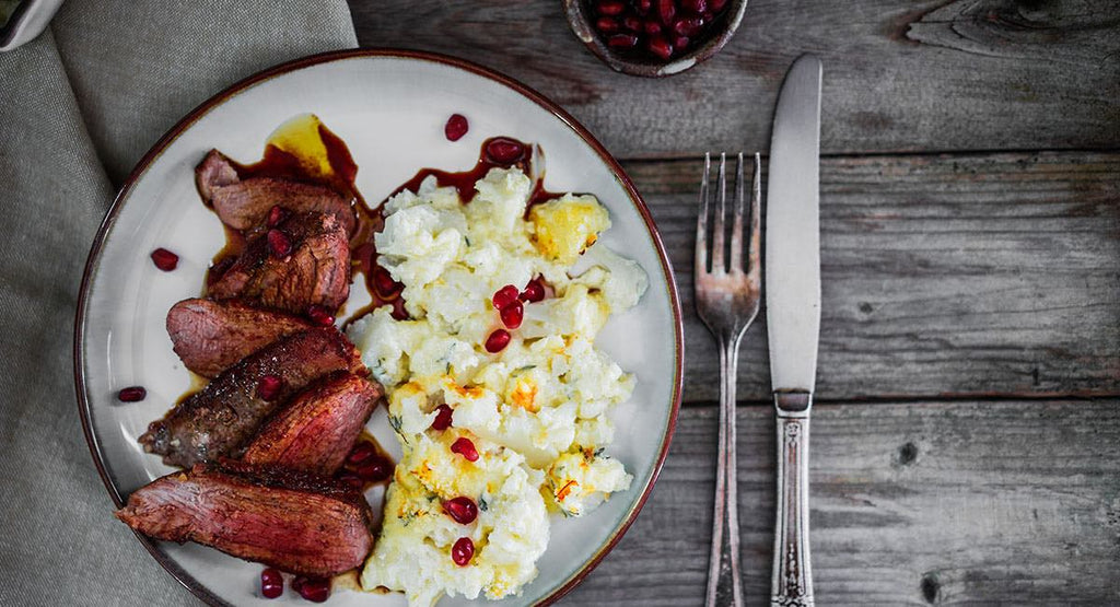 Plate of sliced cooked meat with creamy potatoes topped with pomegranate seeds on a rustic wooden table.