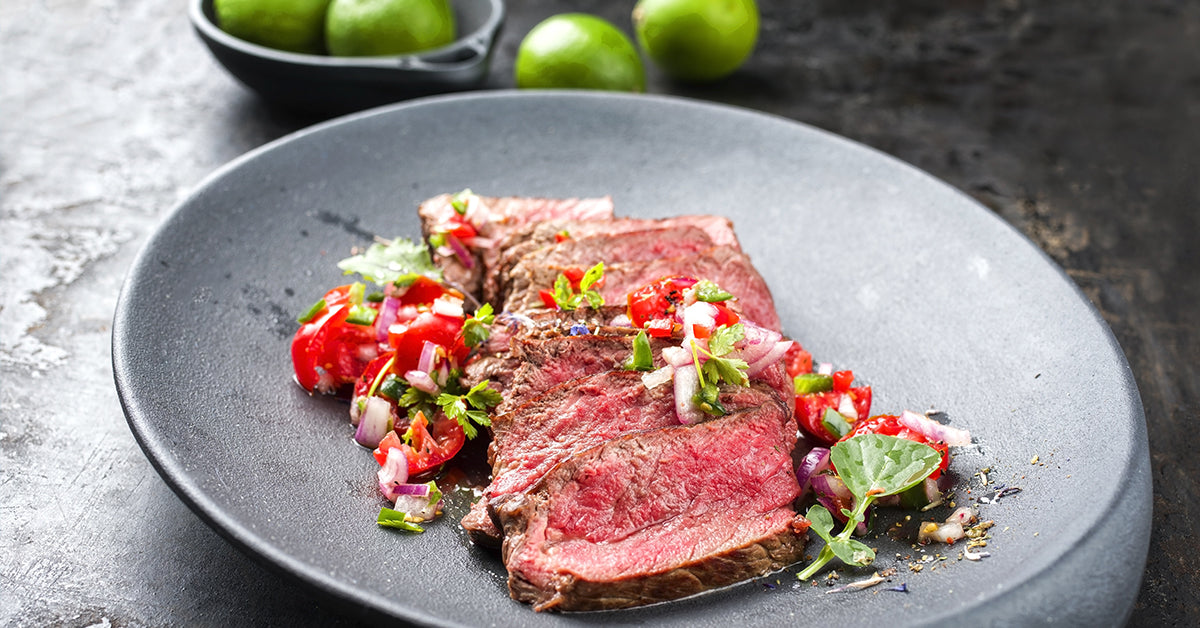 Slices of medium-rare steak topped with fresh tomato and onion salsa on a dark plate.