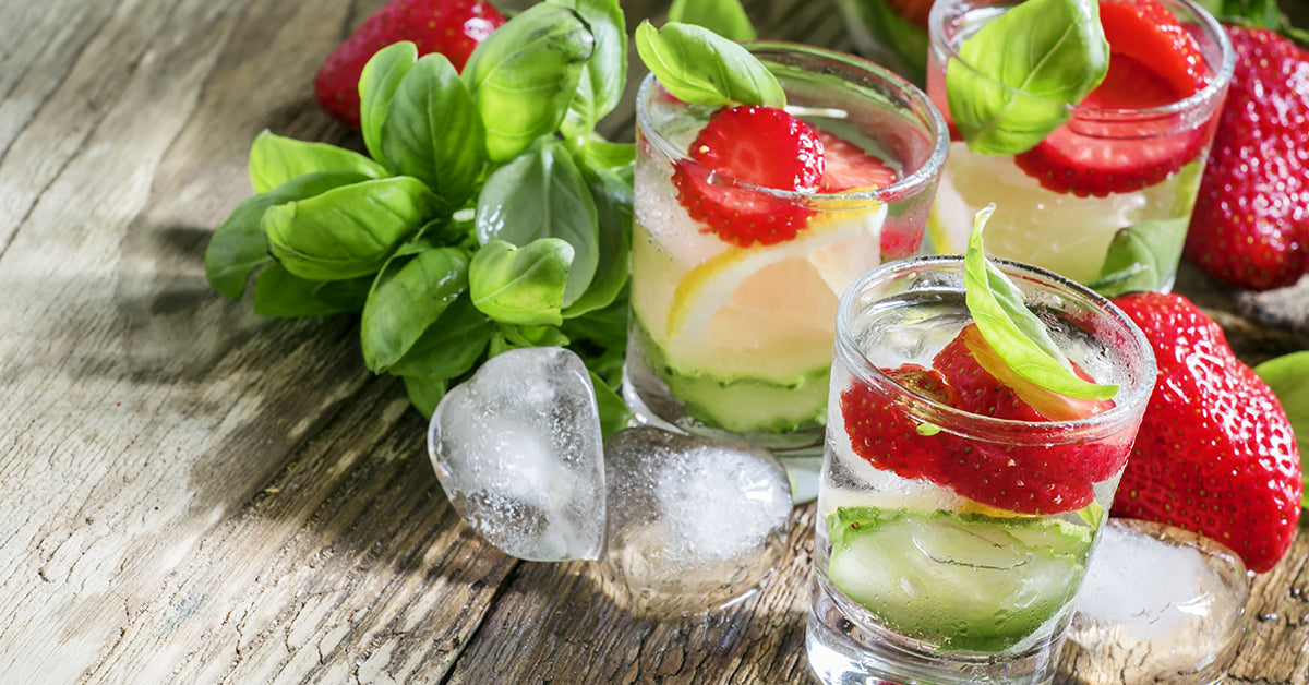 Three clear glasses filled with strawberry and basil-infused detox water sit on a rustic wooden surface, surrounded by fresh strawberries, basil leaves, and ice cubes.