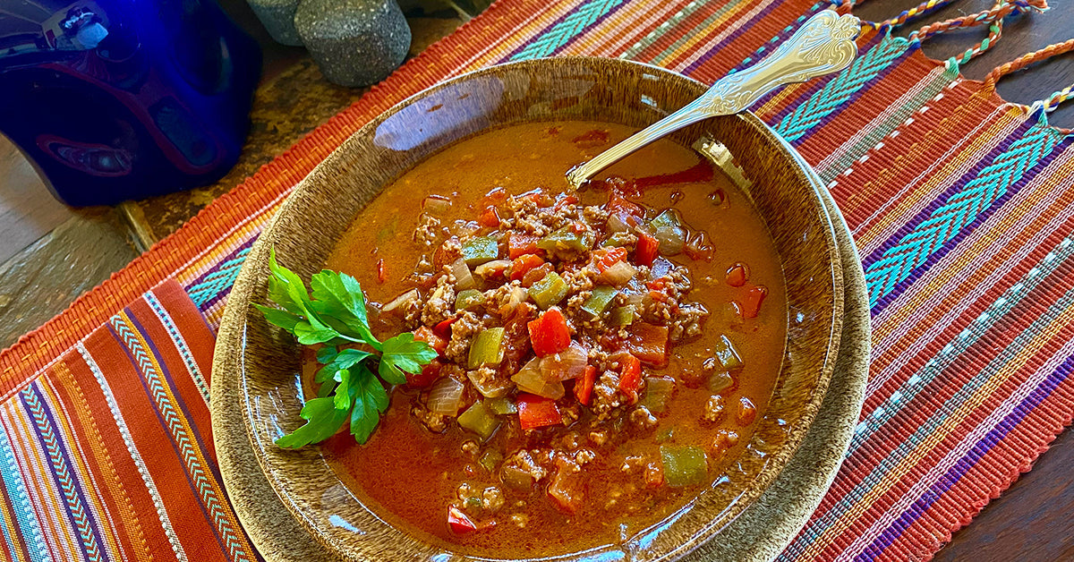 Bowl of hearty stew with ground meat, peppers, onions, and herbs served on a colorful woven tablecloth.