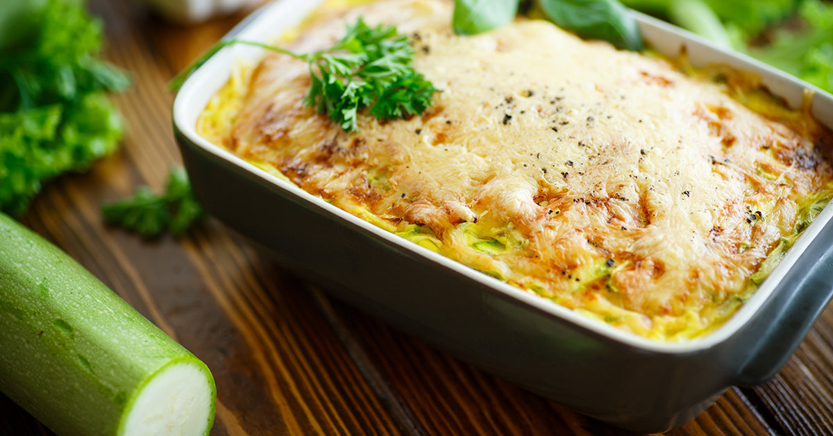 A baked dish with a golden, cheesy top is garnished with fresh herbs, surrounded by fresh vegetables on a wooden table.