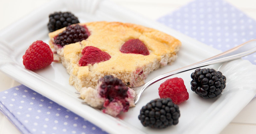 A slice of berry cake topped with raspberries and blackberries is served on a white plate, accompanied by additional berries and a fork, resting on a light blue polka dot napkin.